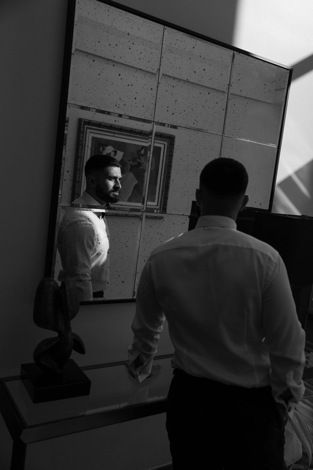 Black and white photo of a bearded man in white shirt viewing his reflection in a large mirror indoors.