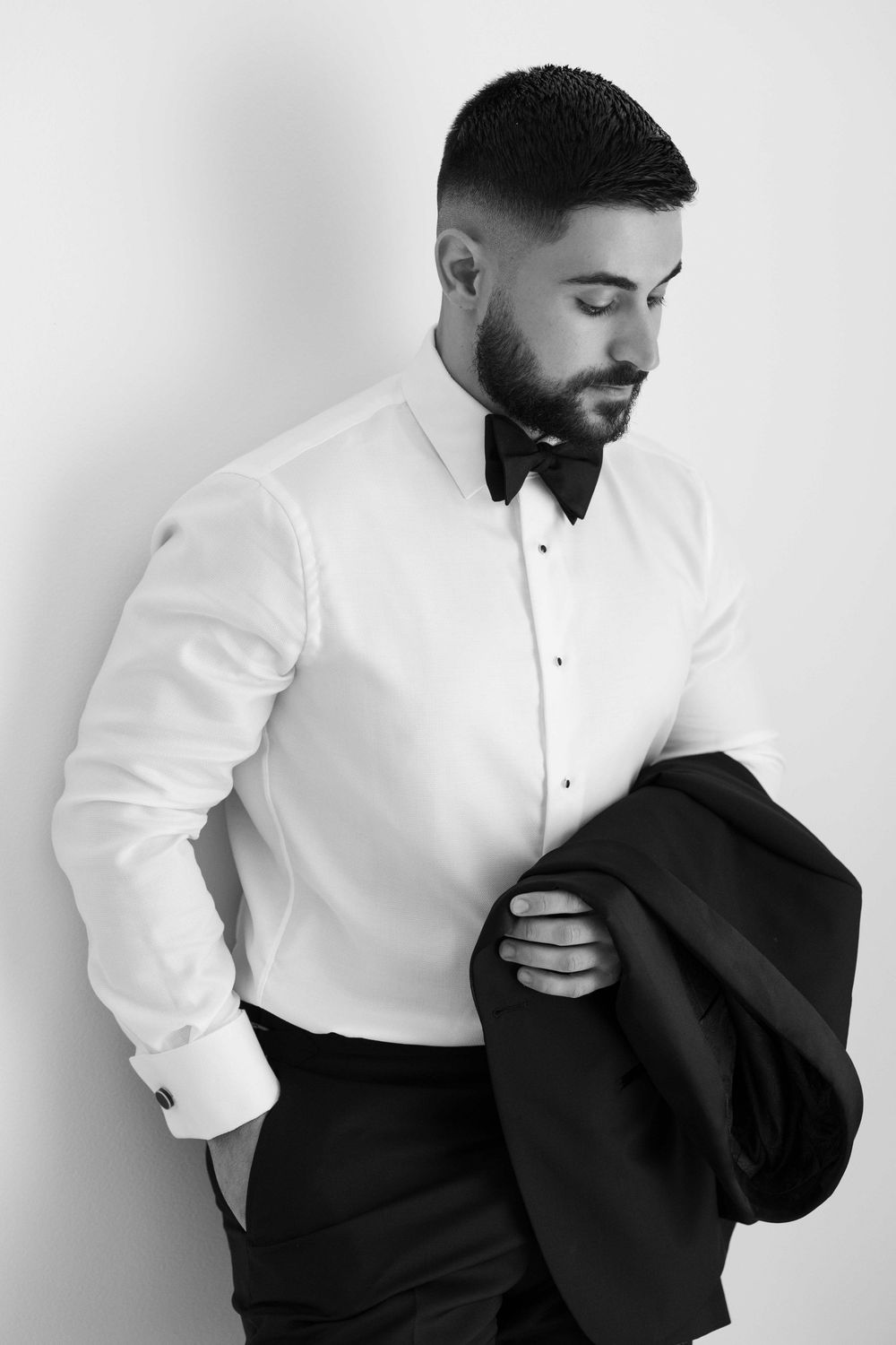 Black and white portrait of a bearded man in a tuxedo shirt and bow tie, holding a suit jacket.
