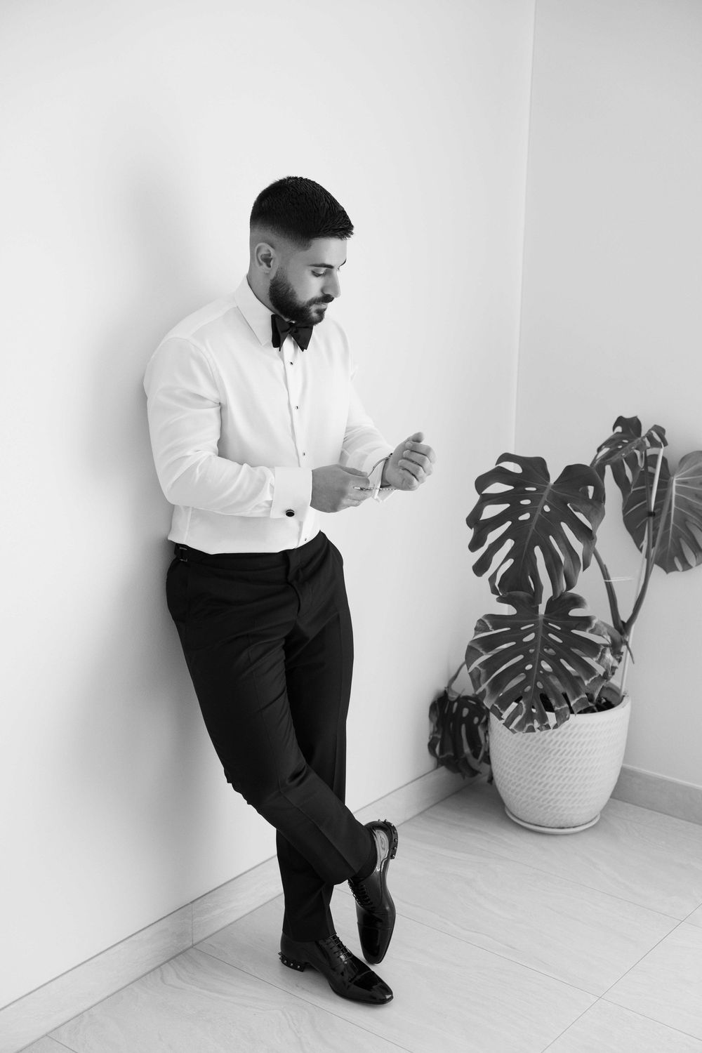 Black and white photo of a well-dressed man in tuxedo shirt and bow tie adjusting cufflinks beside a monstera plant.