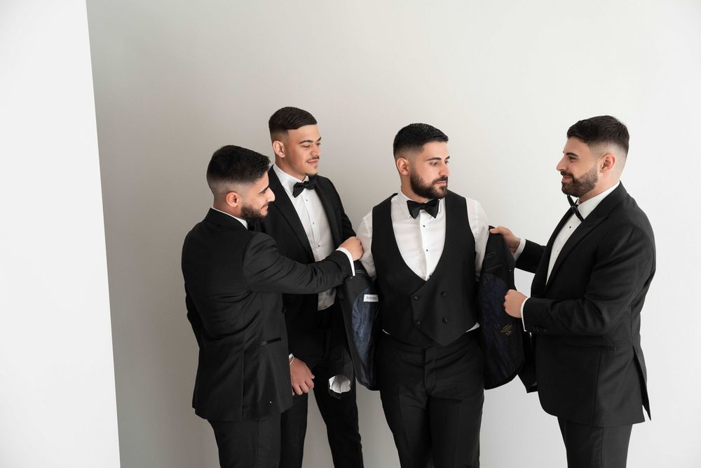 Four men in black tuxedos with bow ties helping each other get dressed before a formal event.