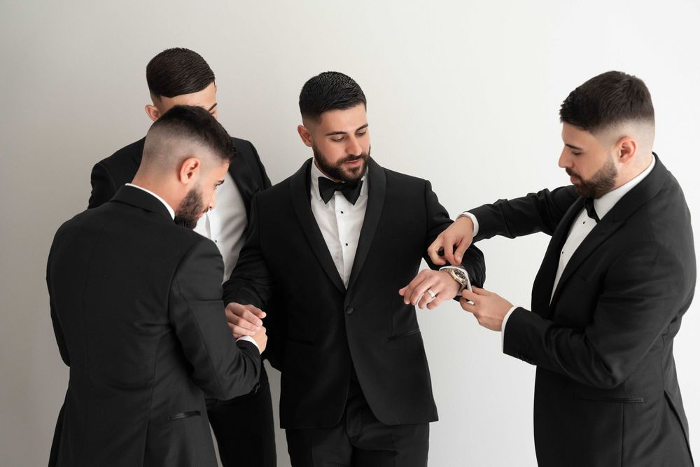 Four men in black tuxedos helping each other with cufflinks and bowties while preparing for a formal event.
