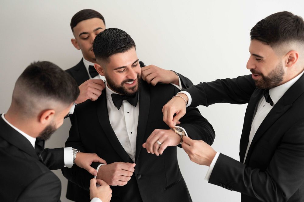 Groomsmen in black tuxedos helping the groom get dressed, adjusting his suit and accessories before the wedding.