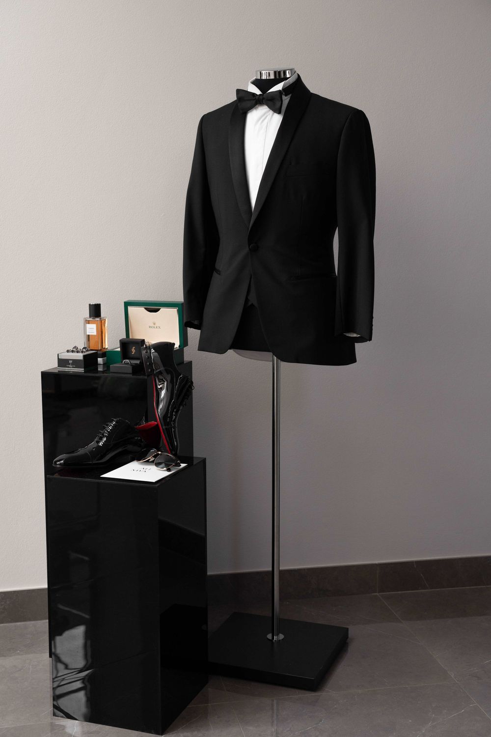 Black tuxedo jacket with bow tie on mannequin beside display of luxury accessories, shoes, and cologne.