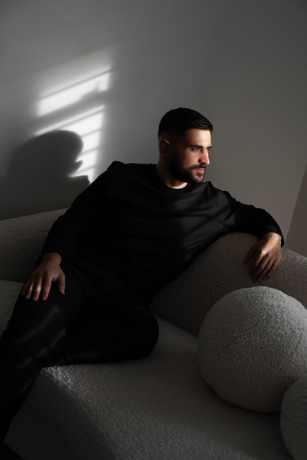 Man in black outfit relaxing on a white textured sofa with dramatic light and shadow on the wall.