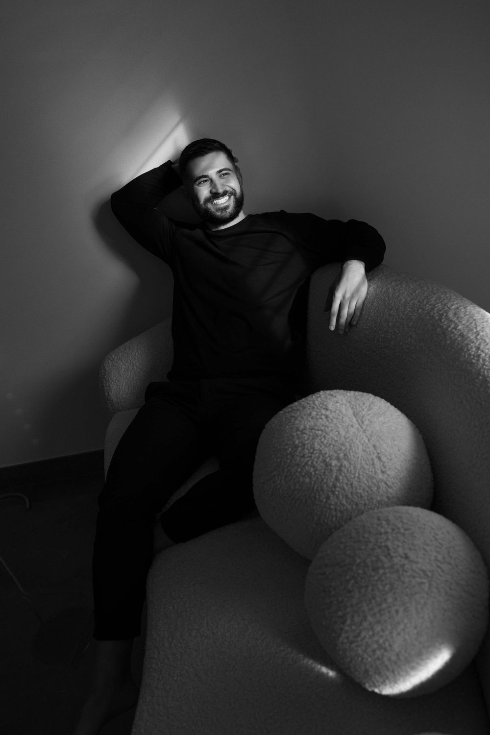 Smiling bearded man in black outfit relaxing on a round fluffy sofa in a moody black and white portrait.