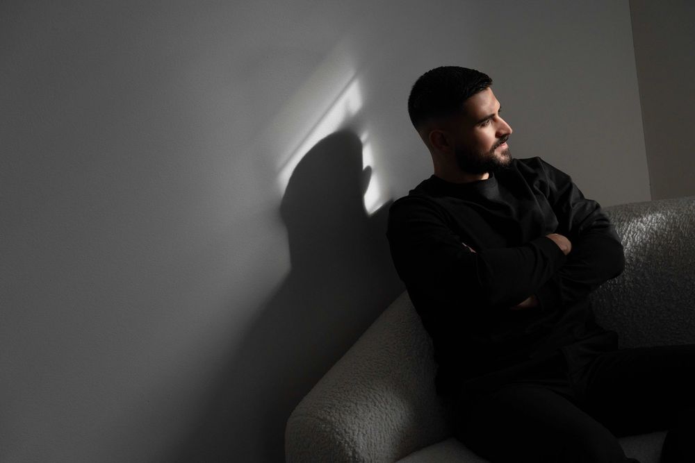 Young man in black sitting on sofa, arms crossed, gazing thoughtfully at wall with dramatic light and shadow.