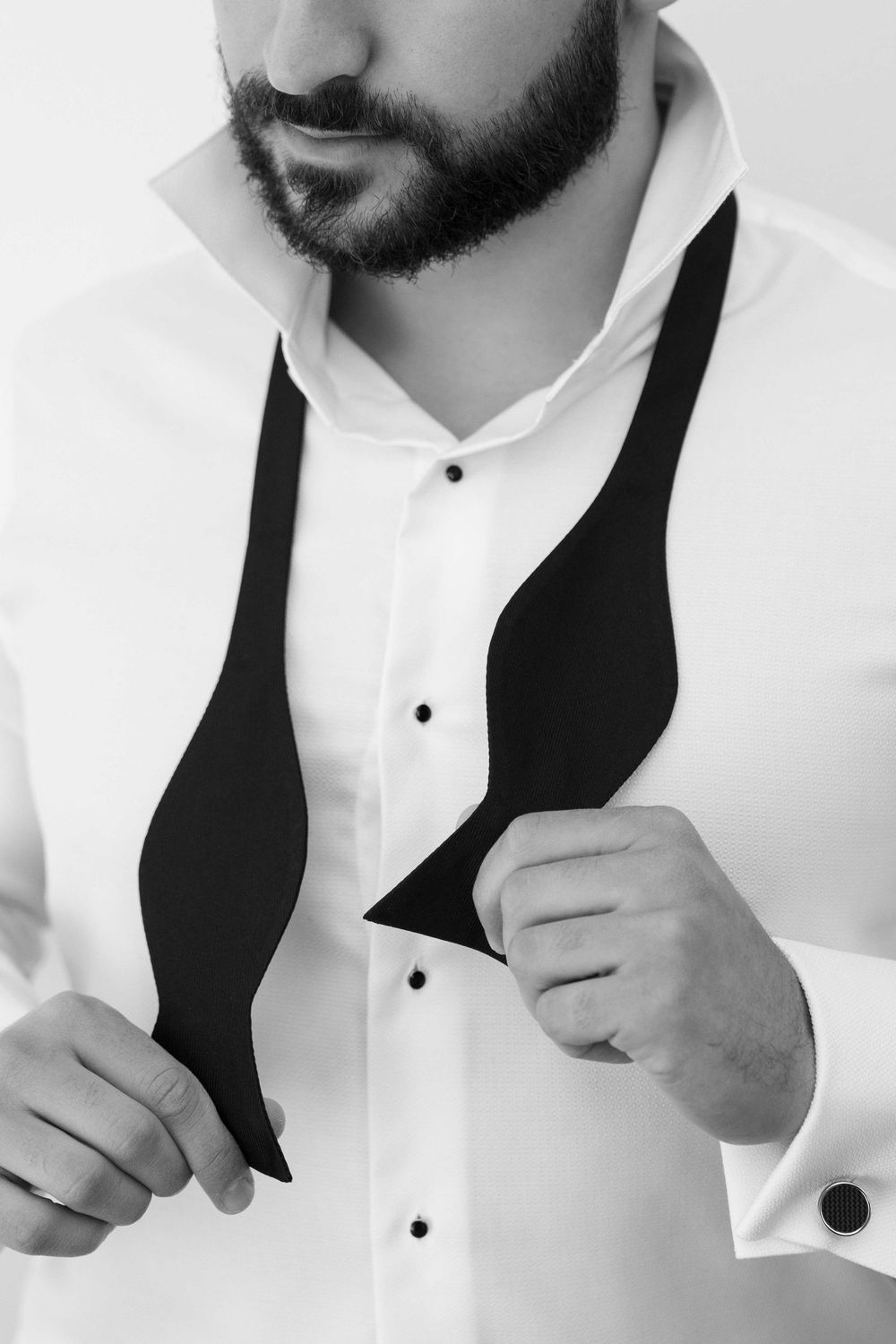 Bearded man in white tuxedo shirt adjusting black bow tie, black and white photo.