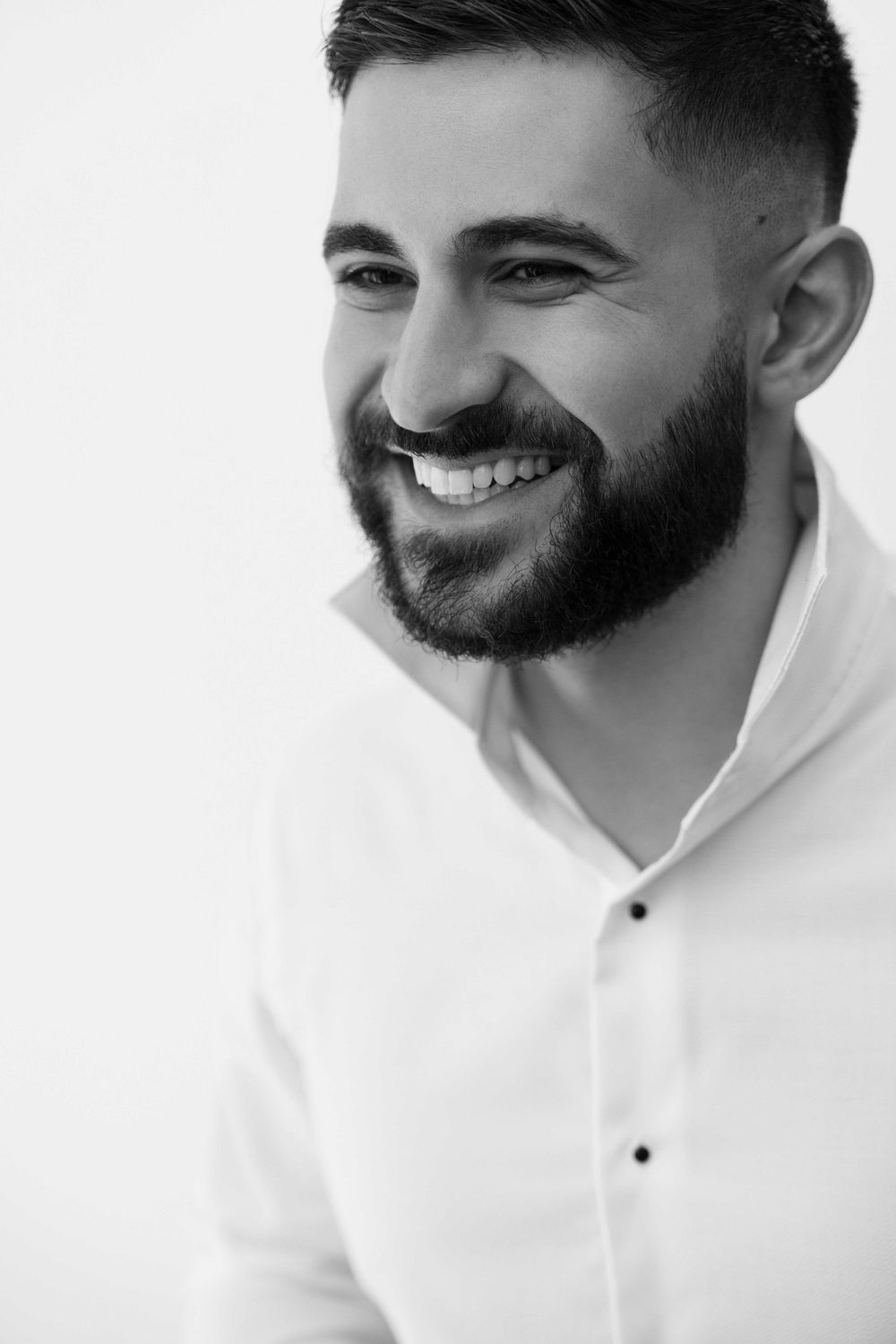 Black and white portrait of a smiling bearded man in a white shirt, exuding confidence and charm.
