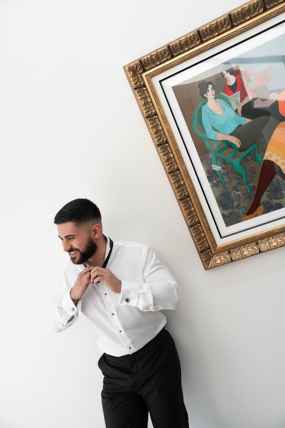 A well-dressed man in a white shirt adjusts his bow tie while standing beside an ornate framed painting on a white wall.