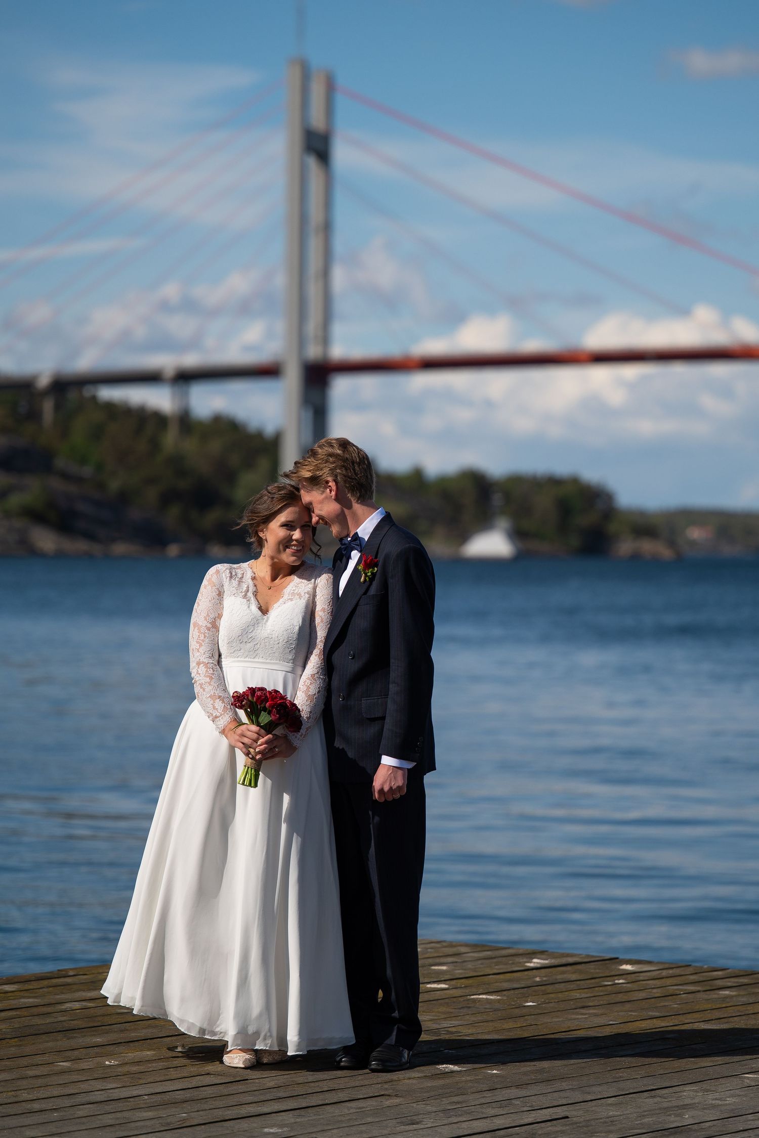 Bride and groom pose on a wooden dock with a cable-stayed bridge and blue water in the background.