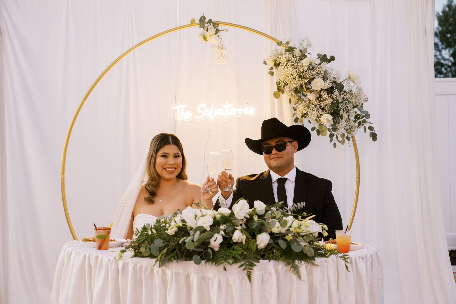 Newlyweds celebrate at their reception table with floral decorations and a neon sign backdrop.