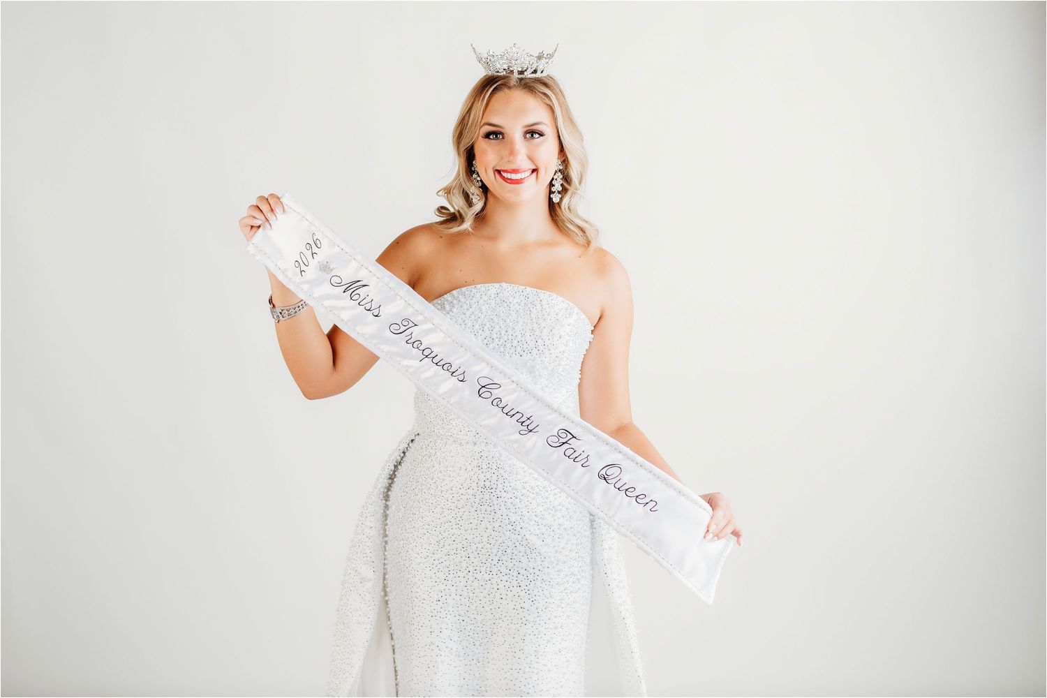 Blonde woman in white sequin gown wearing tiara and holding a pageant sash, posing against white background.