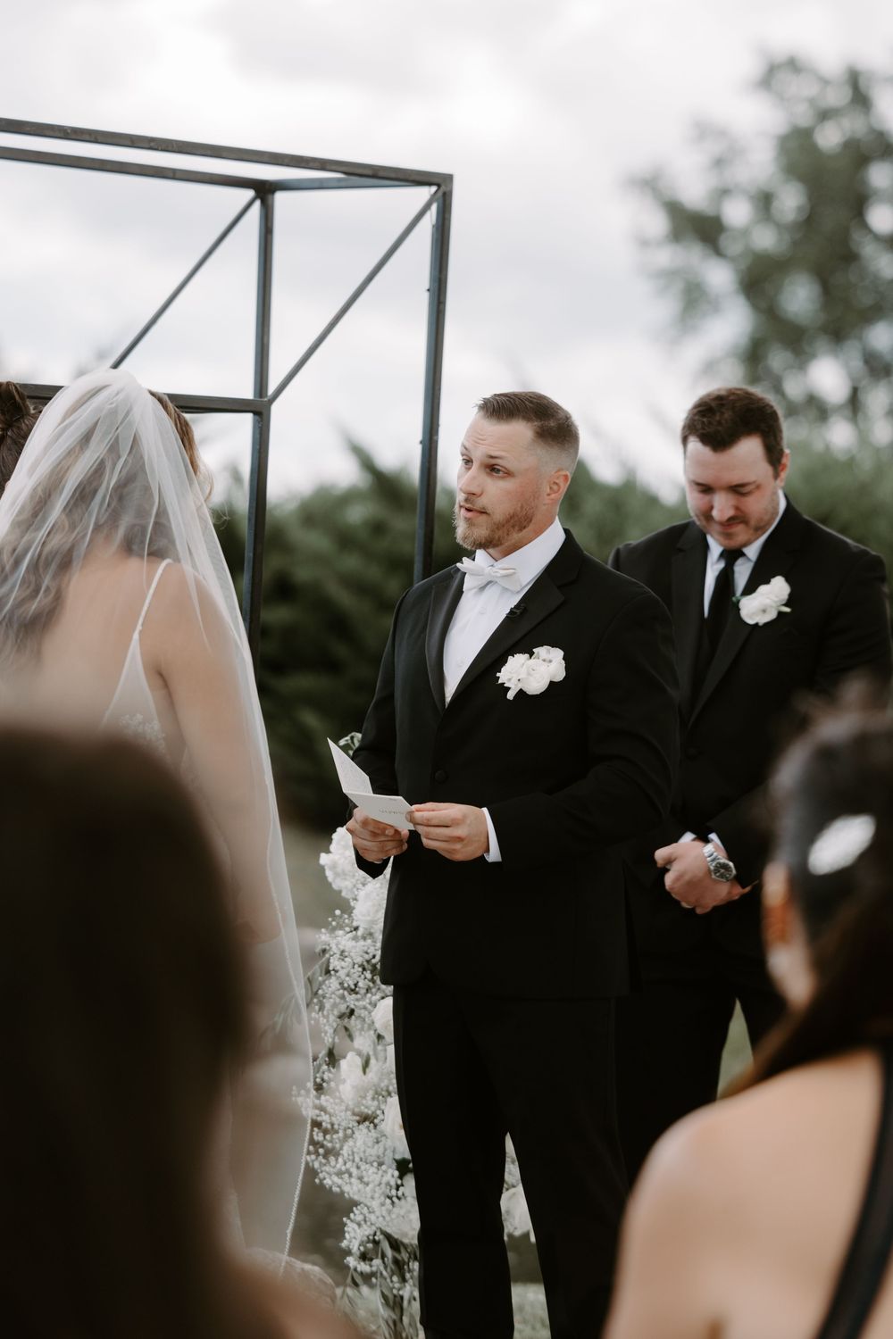 Groom in black tuxedo reads vows to bride at outdoor wedding ceremony beneath geometric arch.