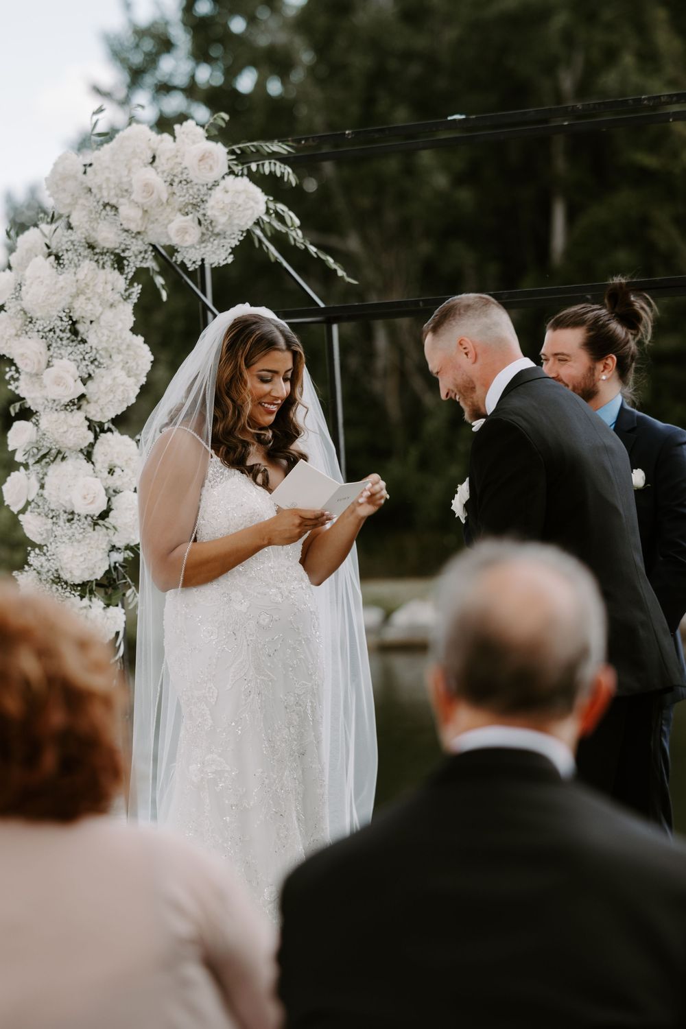 Bride reads vows to groom at outdoor wedding ceremony with white floral arch backdrop.