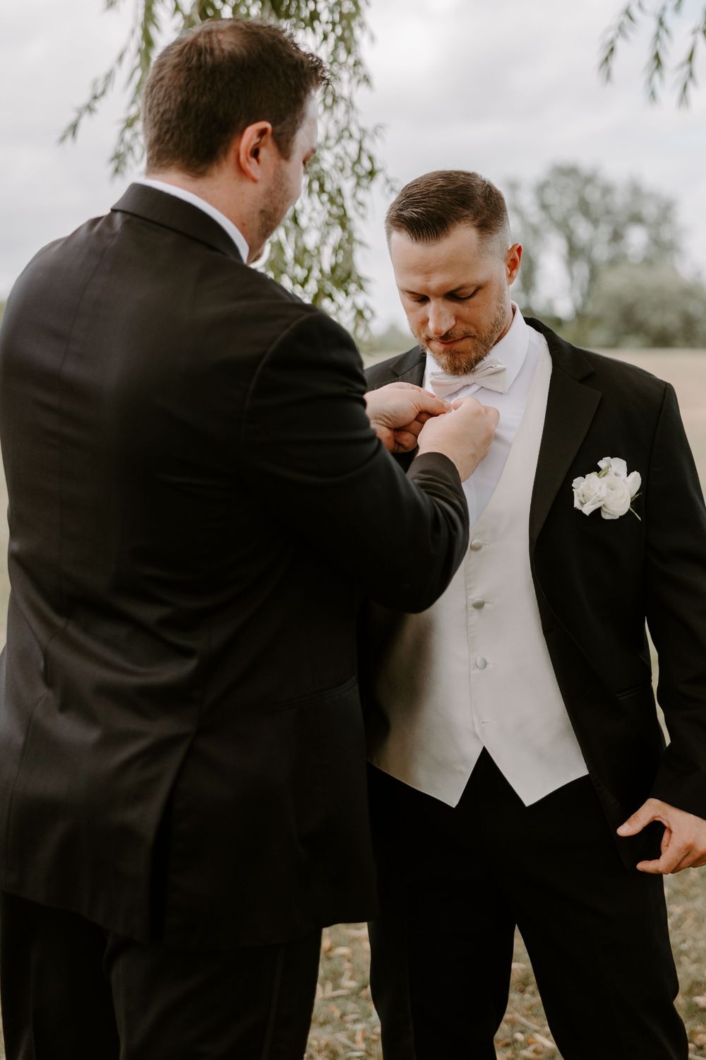 Groomsman adjusts groom's white bow tie before wedding ceremony, both wearing black tuxedos outdoors.