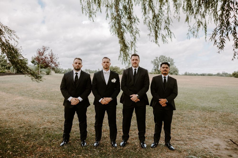 Groom and three groomsmen in black suits pose outdoors under a weeping willow.