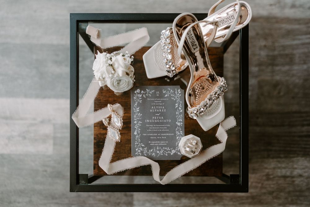Wedding flat lay featuring ornate heeled shoes, floral details, ribbon, and an elegant gray invitation on a glass table.