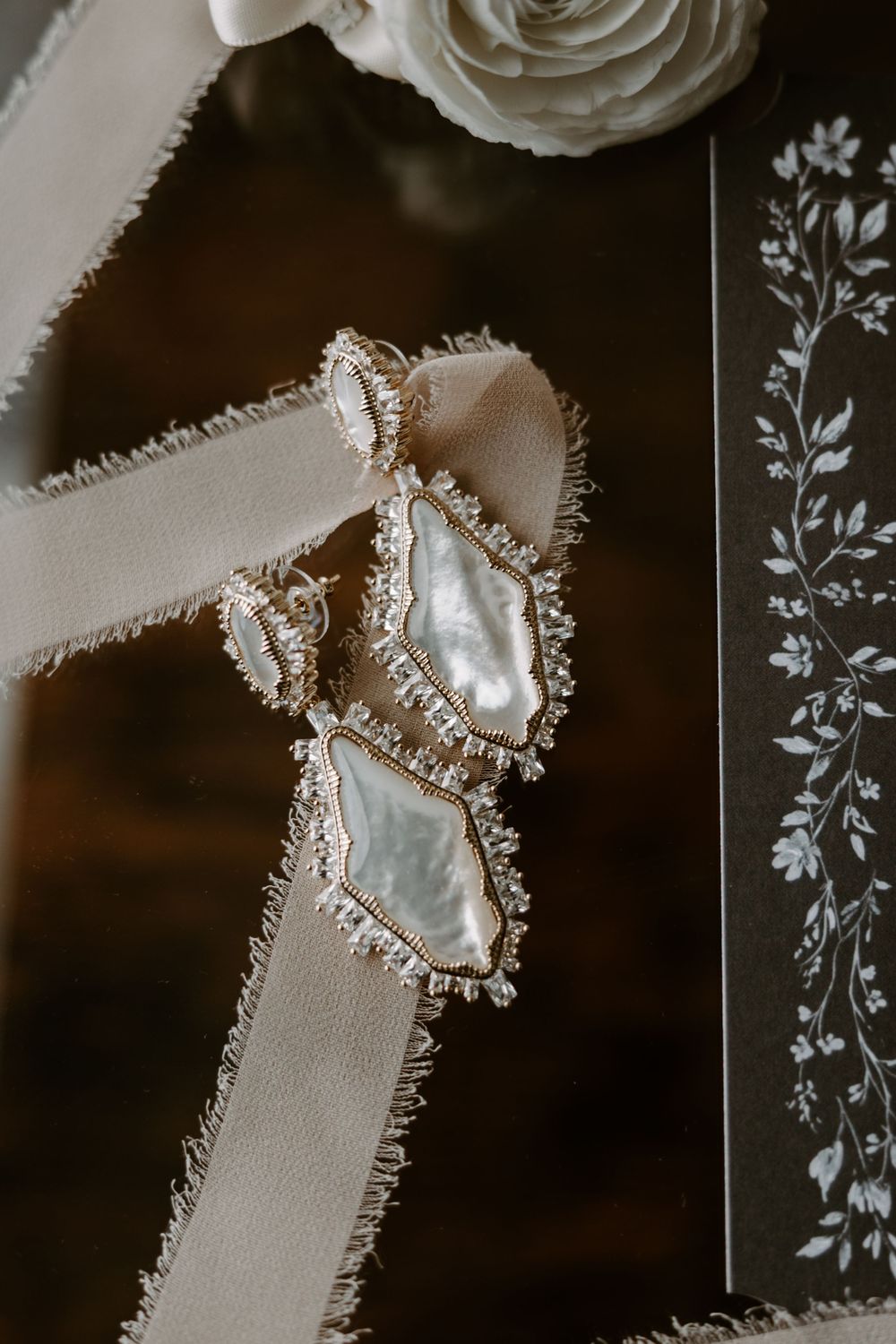 Elegant mother-of-pearl and gold bridal earrings displayed on ribbon beside floral wedding stationery.
