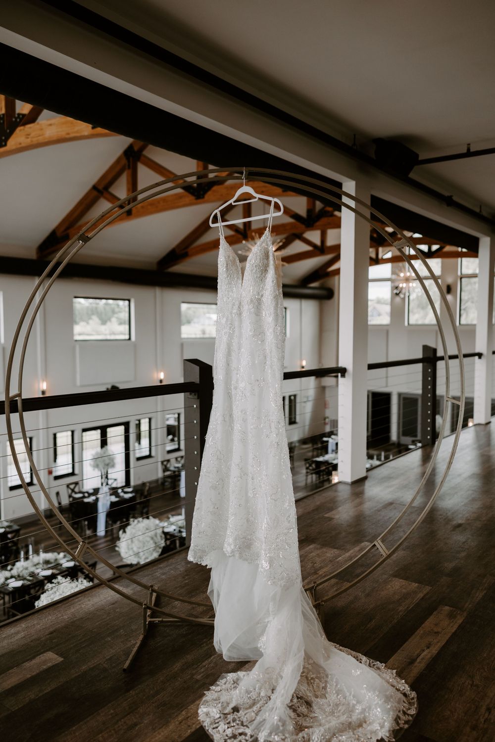 A lace wedding dress hanging from a circular gold arch in an elegant venue with vaulted ceilings and hardwood floors.