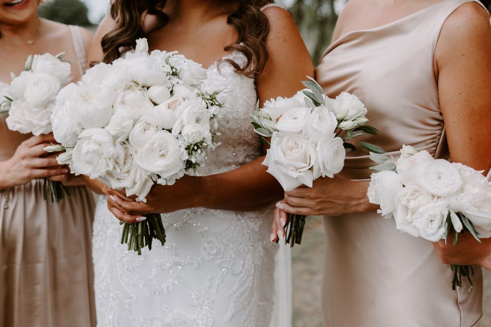 Bride and bridesmaids in champagne dresses holding elegant white rose and peony wedding bouquets.