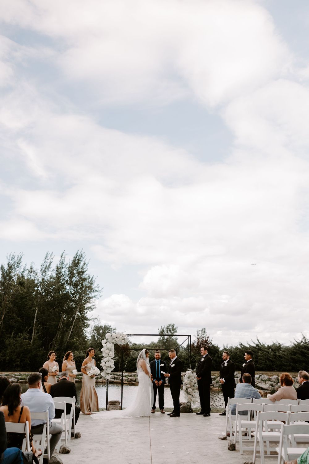 Outdoor wedding ceremony with bride and groom at floral arch altar under dramatic cloudy sky.