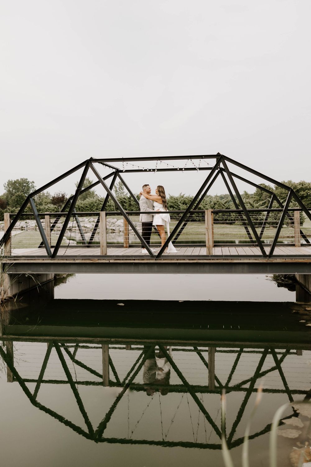 Couple embracing on a black iron bridge reflected in calm water below, surrounded by lush greenery.