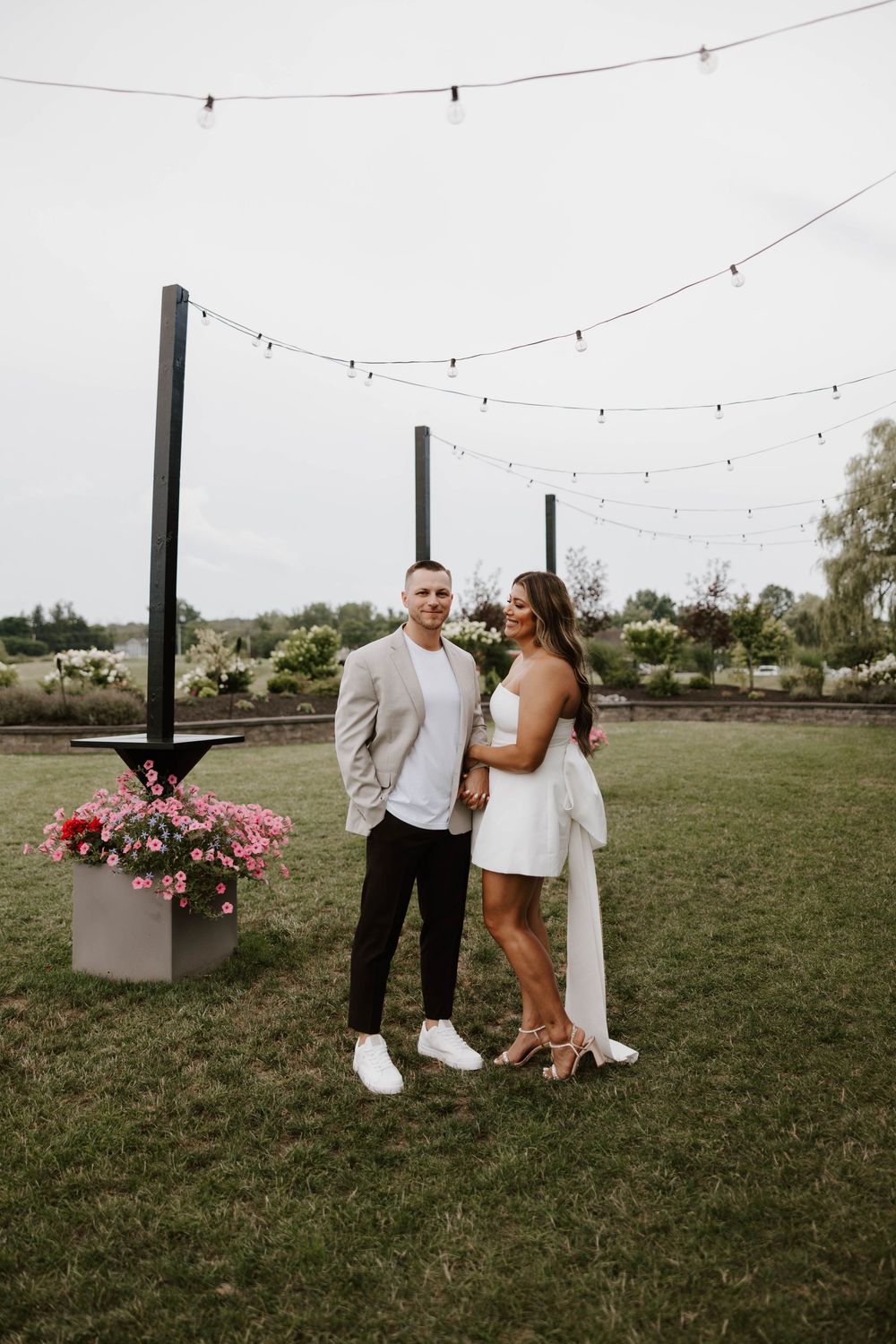 Couple posing outdoors under string lights on green lawn, man in beige blazer, woman in white dress, pink flowers nearby.