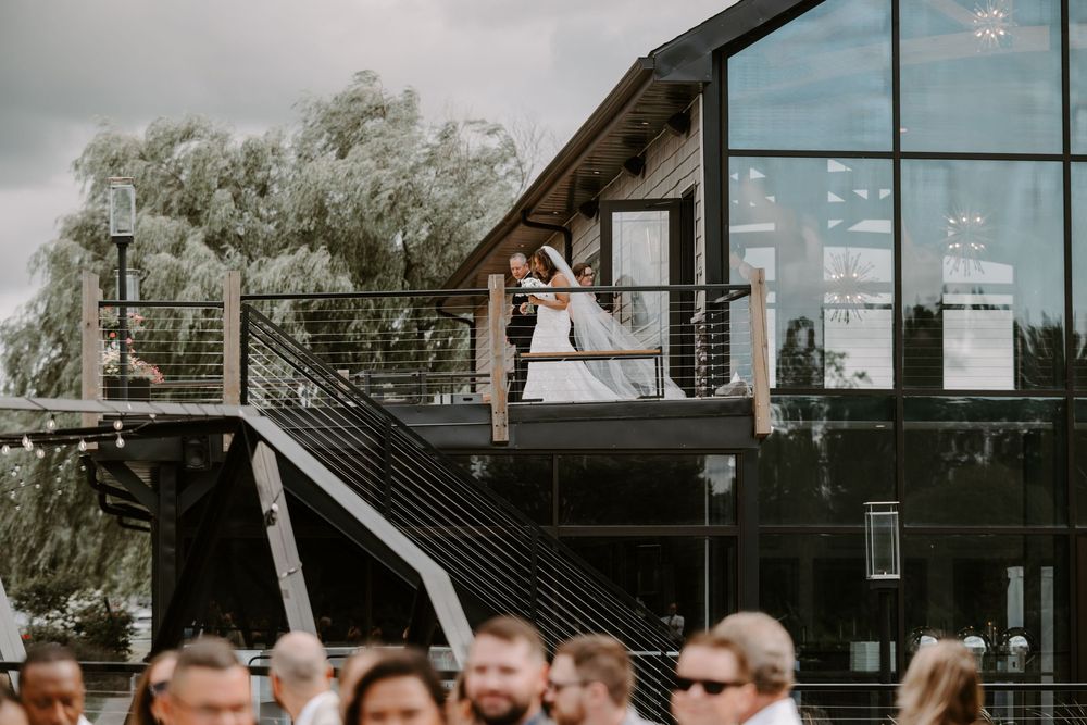 Couple sharing a romantic kiss on a modern venue balcony during an outdoor wedding ceremony with guests below.