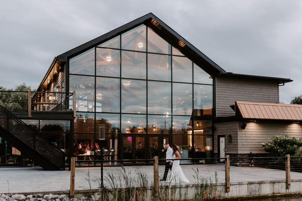 Bride and groom walk outside a stunning glass barn wedding venue at dusk with warm interior lights glowing.