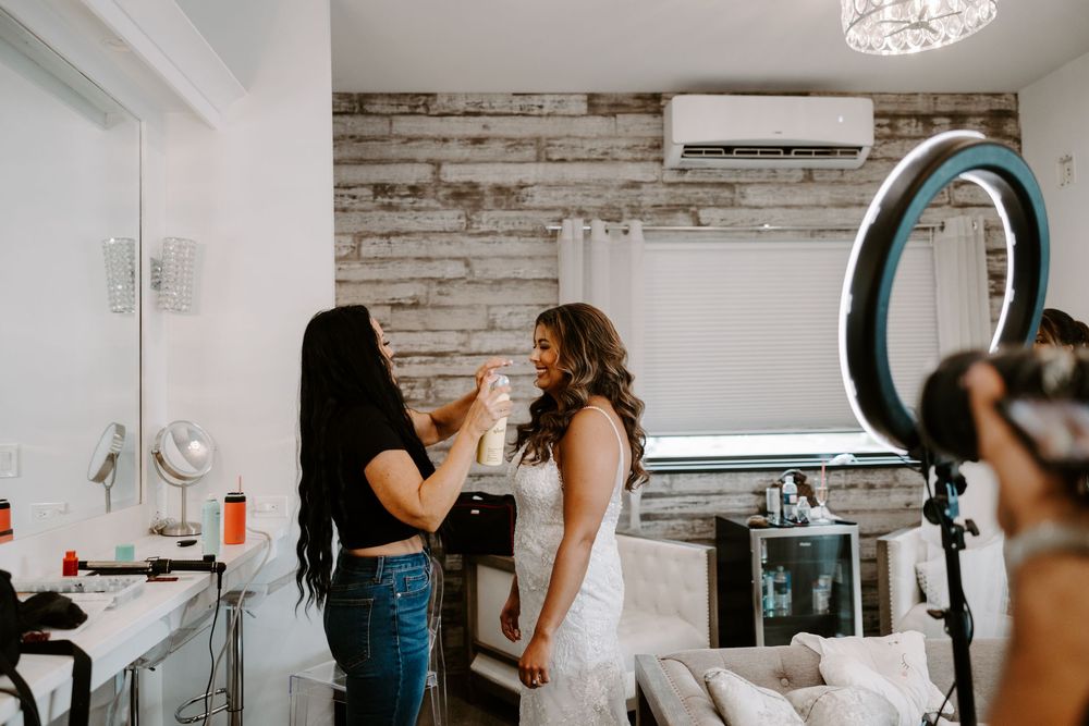 Makeup artist applying finishing touches to a bride in a white wedding dress inside a bright modern bridal suite.