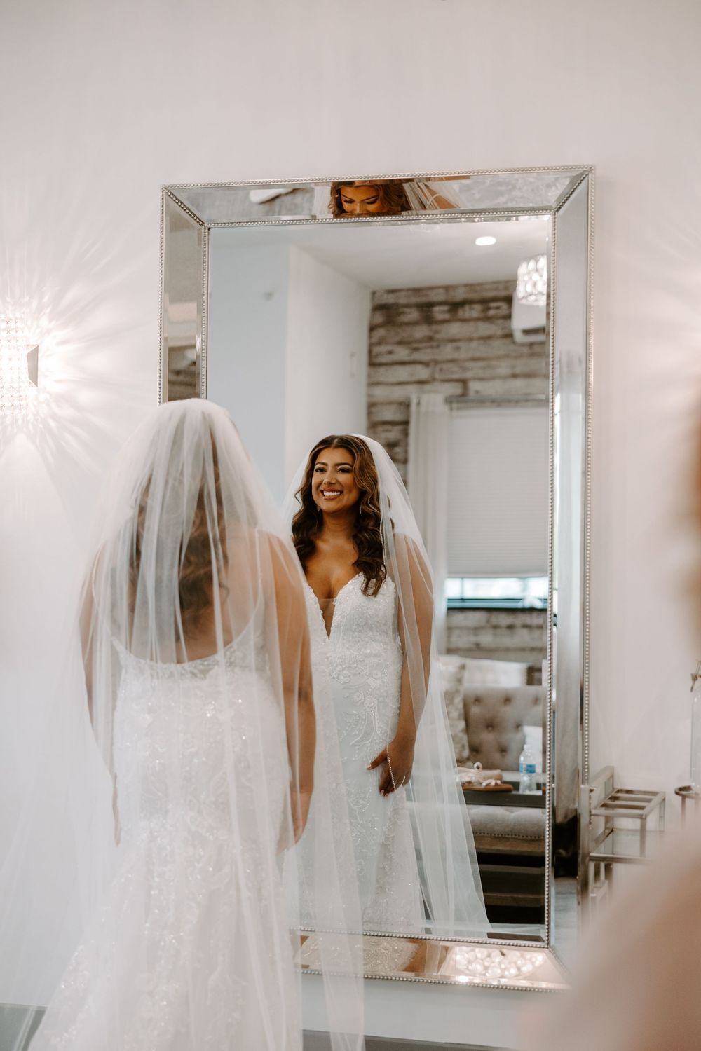 Smiling bride in white lace gown and veil admires her reflection in a large mirror on her wedding day.