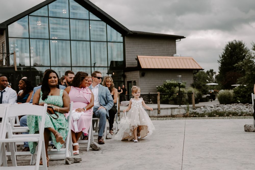 A flower girl walks down the aisle at an outdoor wedding ceremony with guests seated in white chairs.