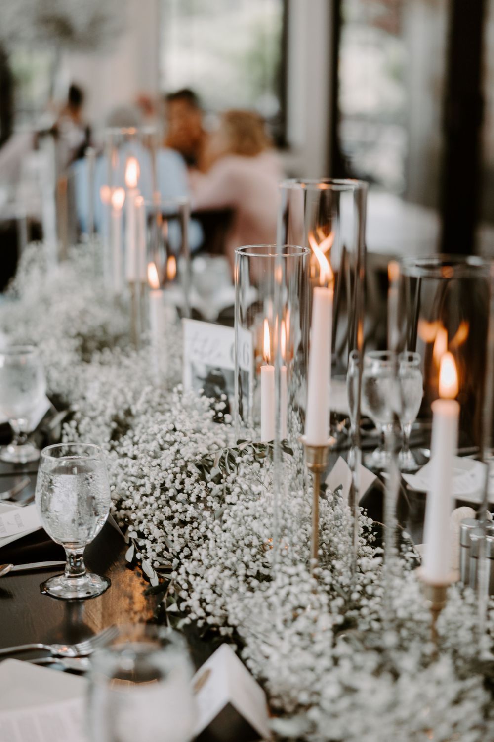 Elegant wedding reception table with white baby's breath, tall candles in glass holders, and crystal glassware on dark table.