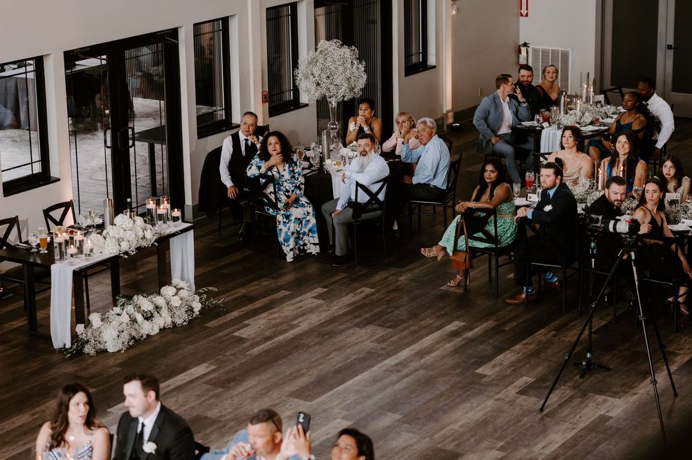 Wedding reception guests seated at decorated tables in a modern venue with dark hardwood floors and floral centerpieces.