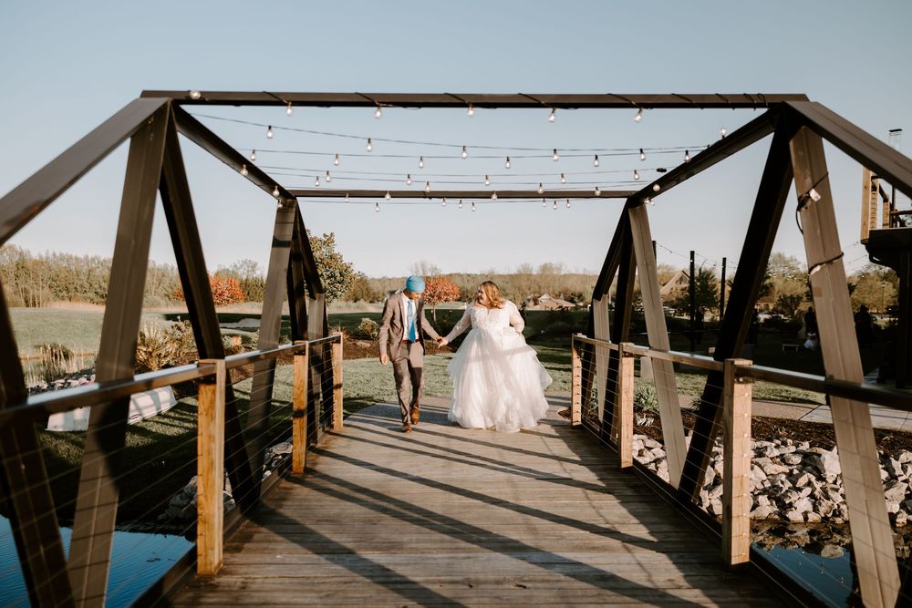 Bride and groom holding hands walking across a wooden bridge decorated with string lights at an outdoor wedding venue.