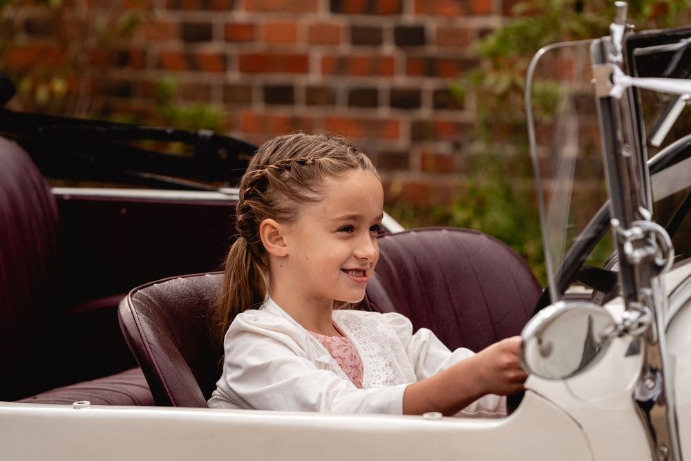 A smiling young girl with braided hair sits in the driver's seat of a vintage white convertible car.