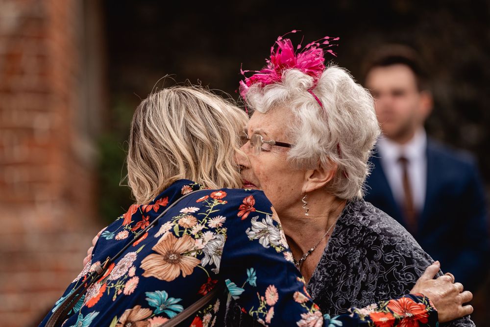 Two women sharing a warm embrace and kiss at a wedding, one wearing a pink floral fascinator.
