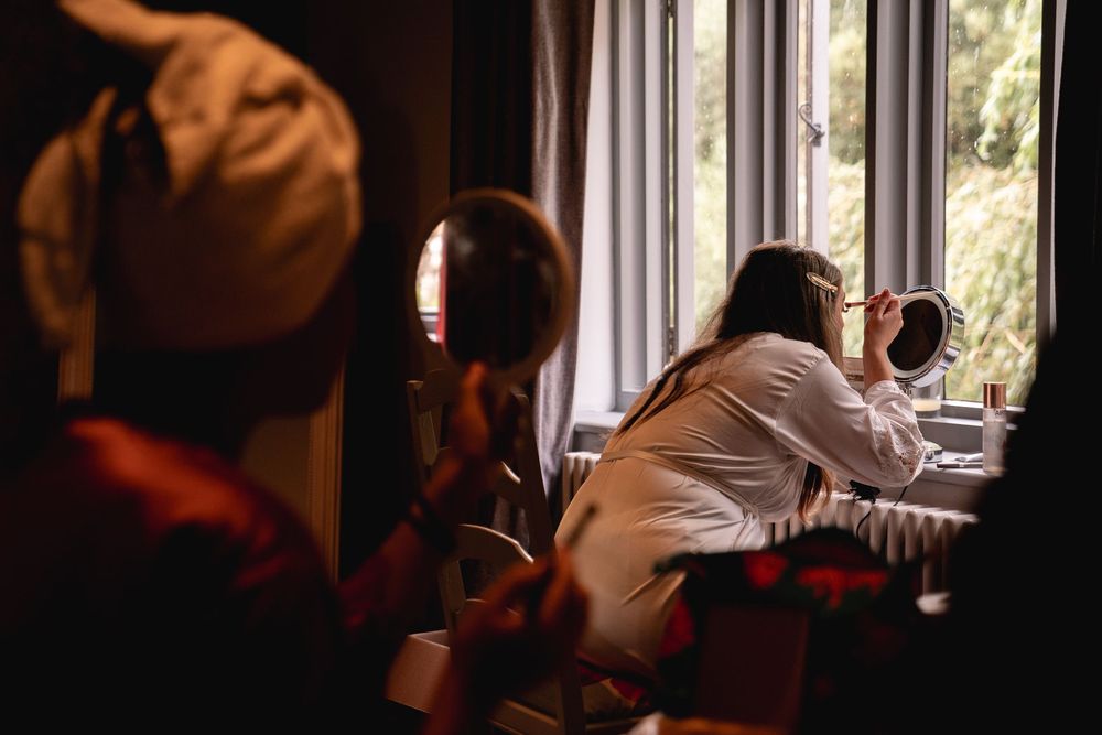 Bride in white robe applying makeup by a window during wedding morning preparations.