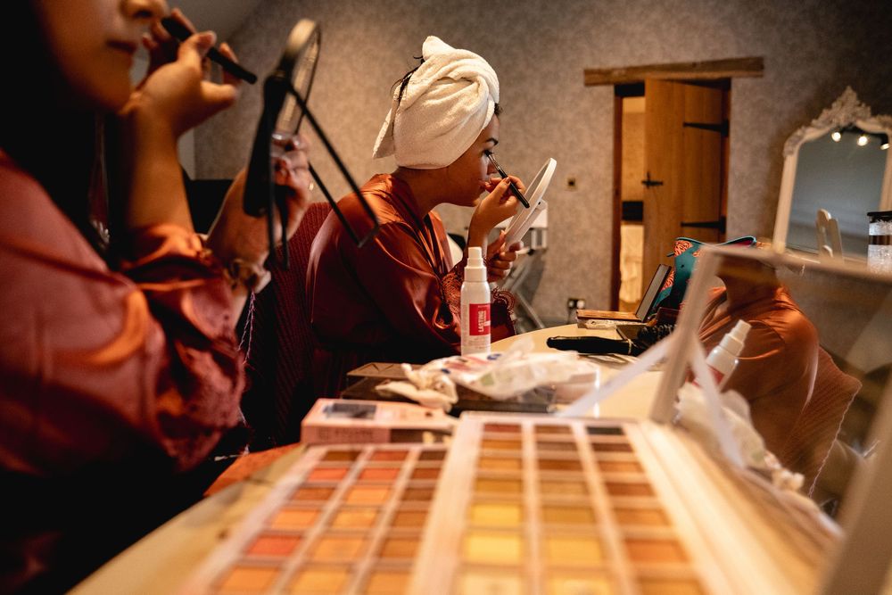 Bridesmaids apply makeup at a vanity with colorful eyeshadow palettes before a wedding.