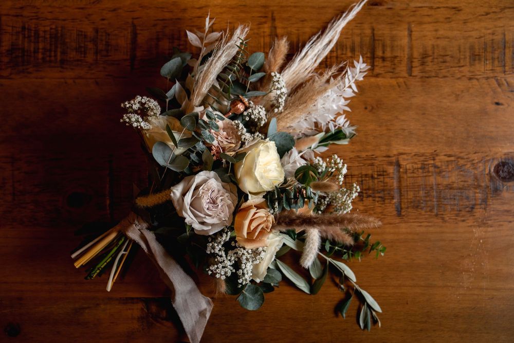 Rustic wedding bouquet with blush roses, pampas grass, eucalyptus, and dried flowers on a wooden surface.