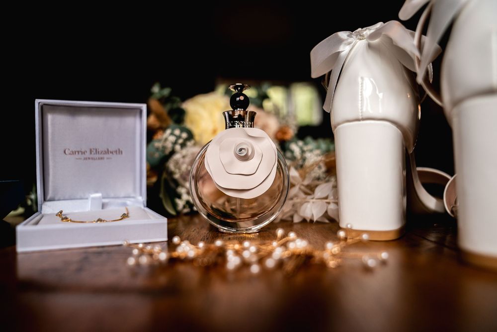 Bridal accessories flat lay featuring white heels, perfume bottle, jewelry box, and pearl necklace on wooden table.