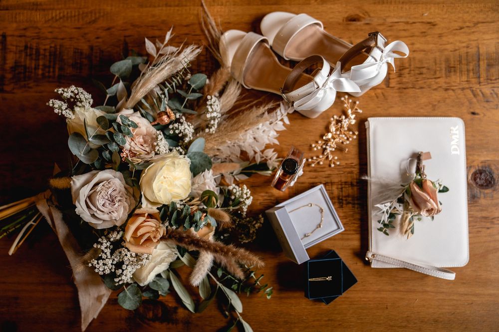 Flat lay of wedding details including a floral bouquet, heels, rings, and boutonniere on a wooden table.