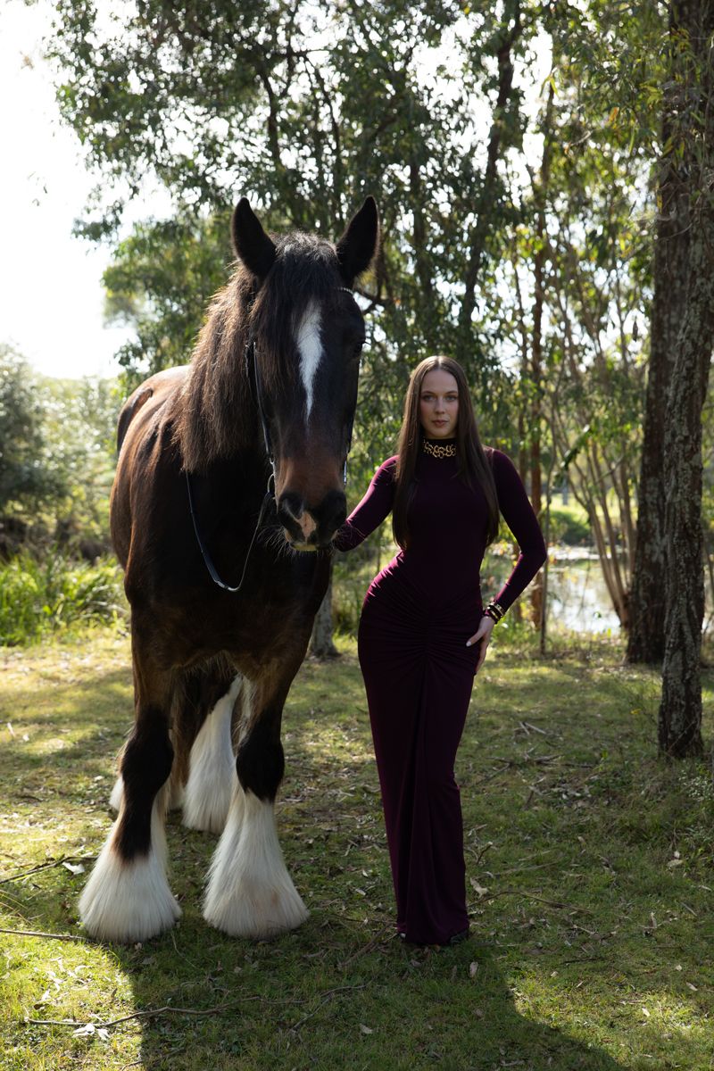 Woman in burgundy jumpsuit stands beside a majestic shire horse in a lush green forest setting during a photoshoot with a horse in sydney