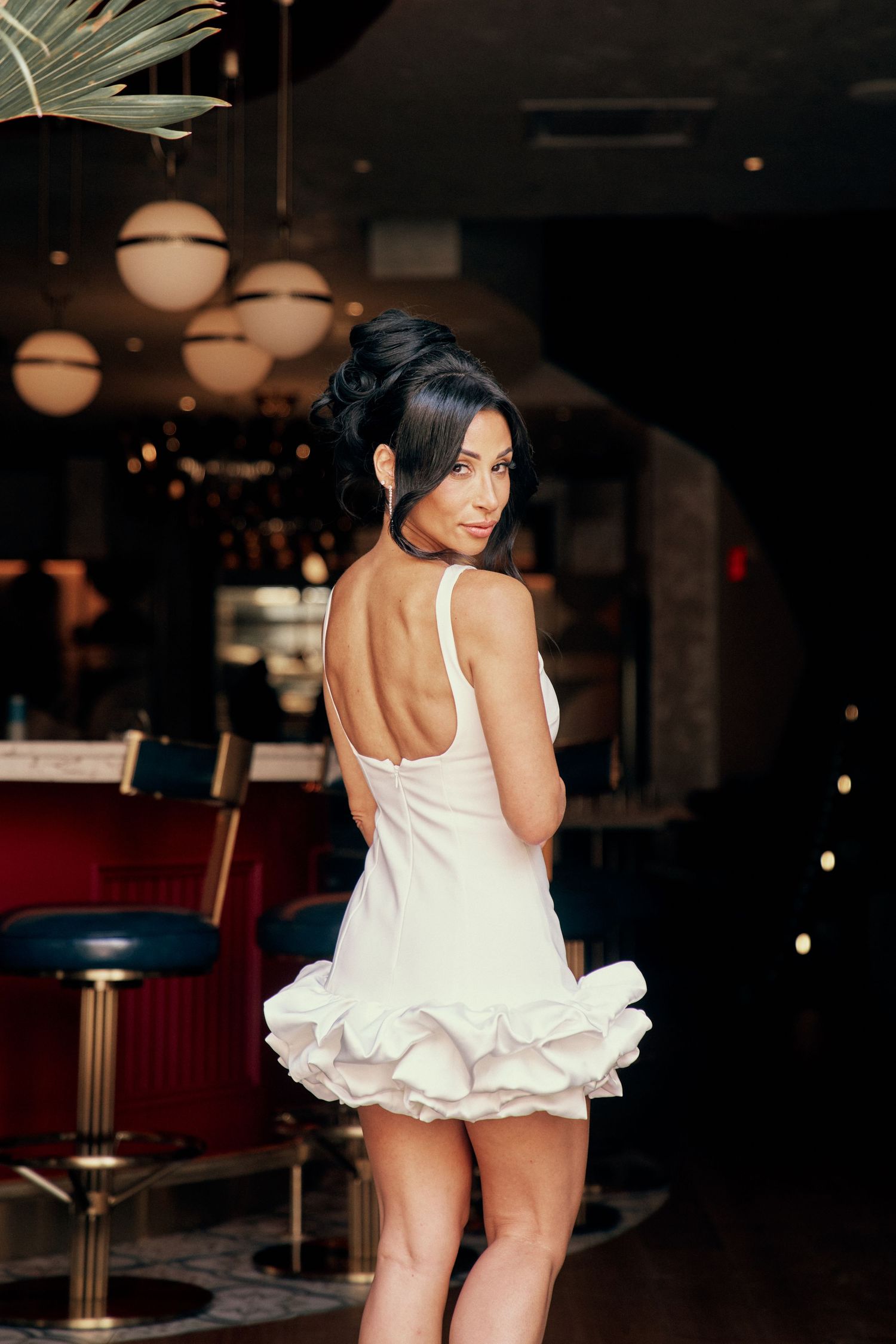 Woman in white backless ruffled mini dress posing in elegant bar setting with warm lighting.