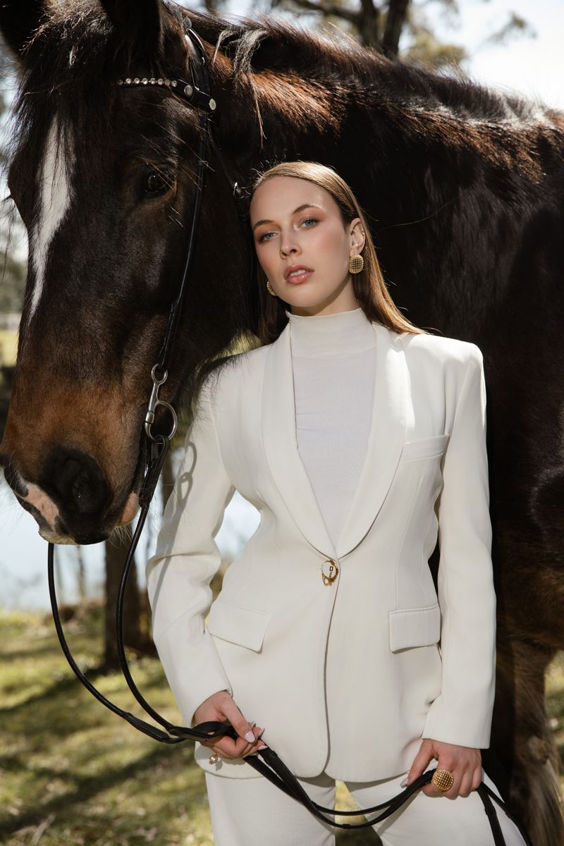 Woman in white blazer suit holding horse reins beside a dark brown horse outdoors during a photoshoot in sydney with a horse