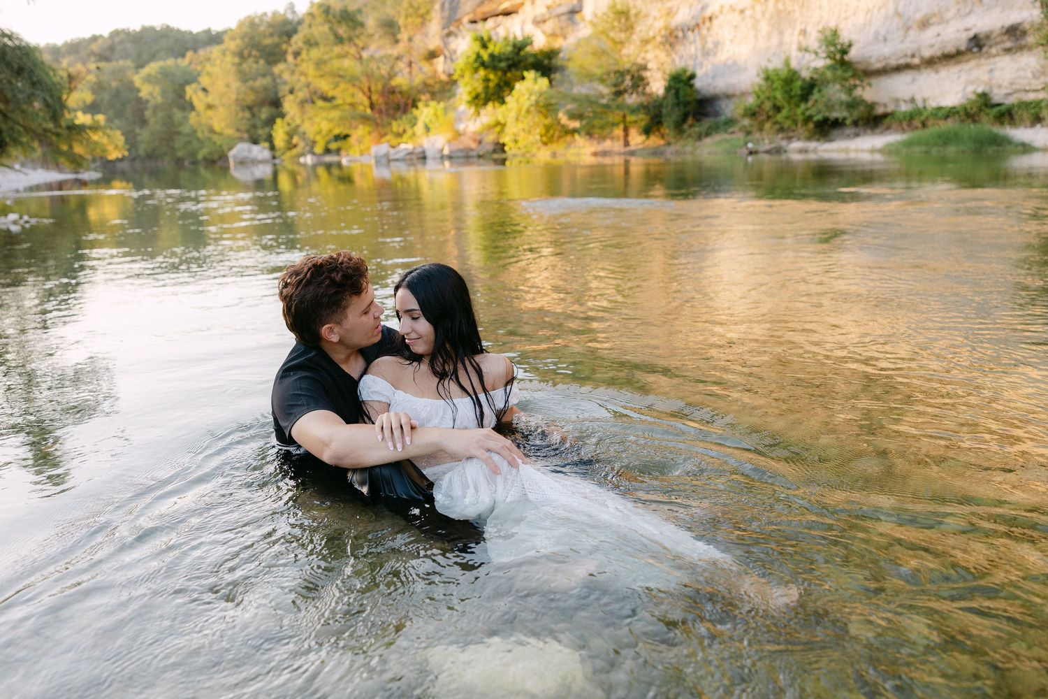 Couple embracing romantically in a shallow river surrounded by lush green trees and rocky cliffs.