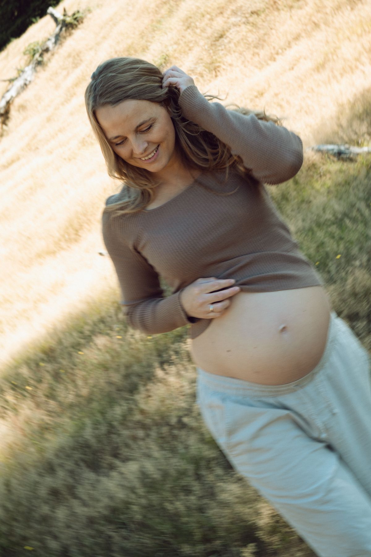 Smiling pregnant woman in brown top and gray pants holds her baby bump outdoors in a sunlit grassy field.