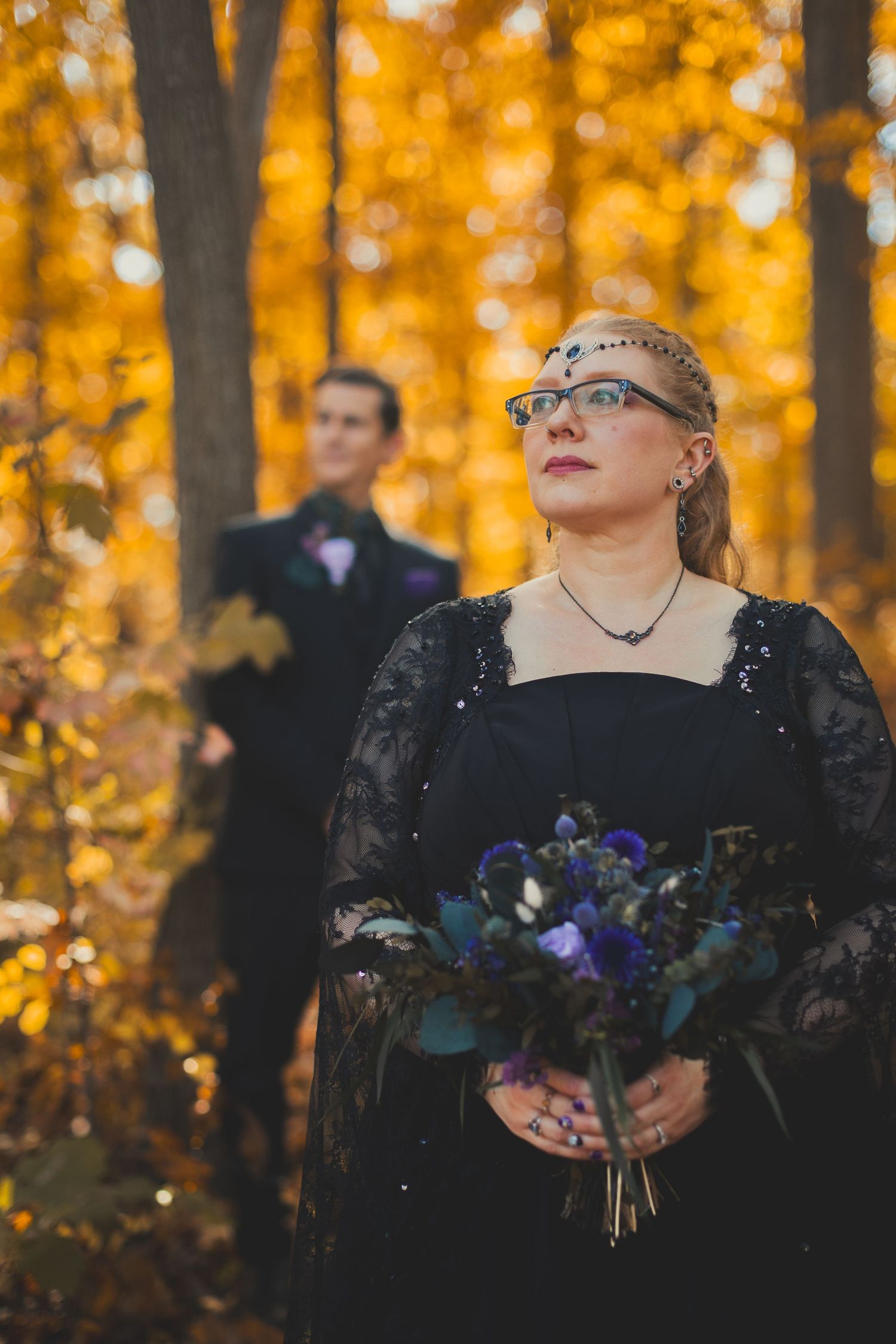 Bride in black lace gown holds purple bouquet in autumn forest, groom in dark suit stands behind among golden trees.