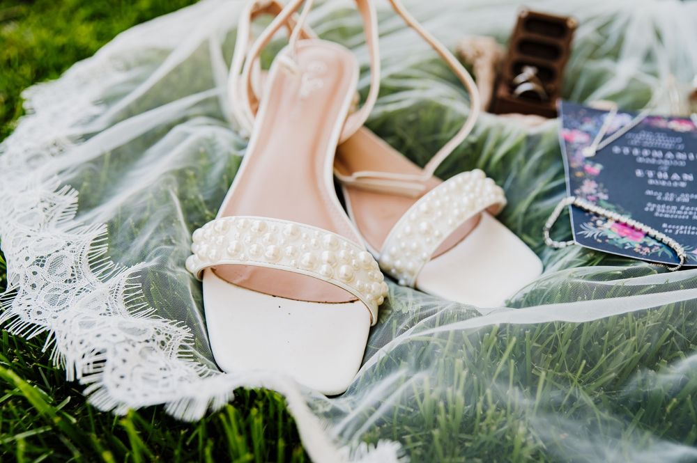 White pearl-studded bridal heels resting on a lace veil beside a floral wedding invitation on green grass.