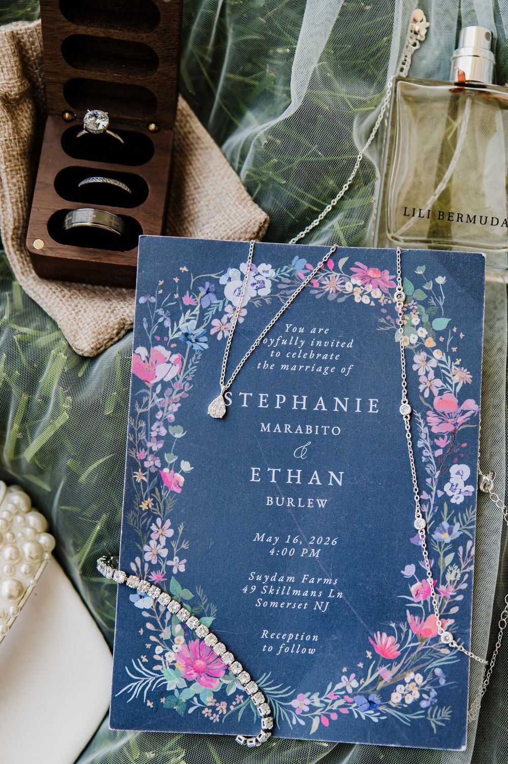 Navy floral wedding invitation for Stephanie Marabito and Ethan Burlew, surrounded by jewelry, rings, and perfume on green fabric.